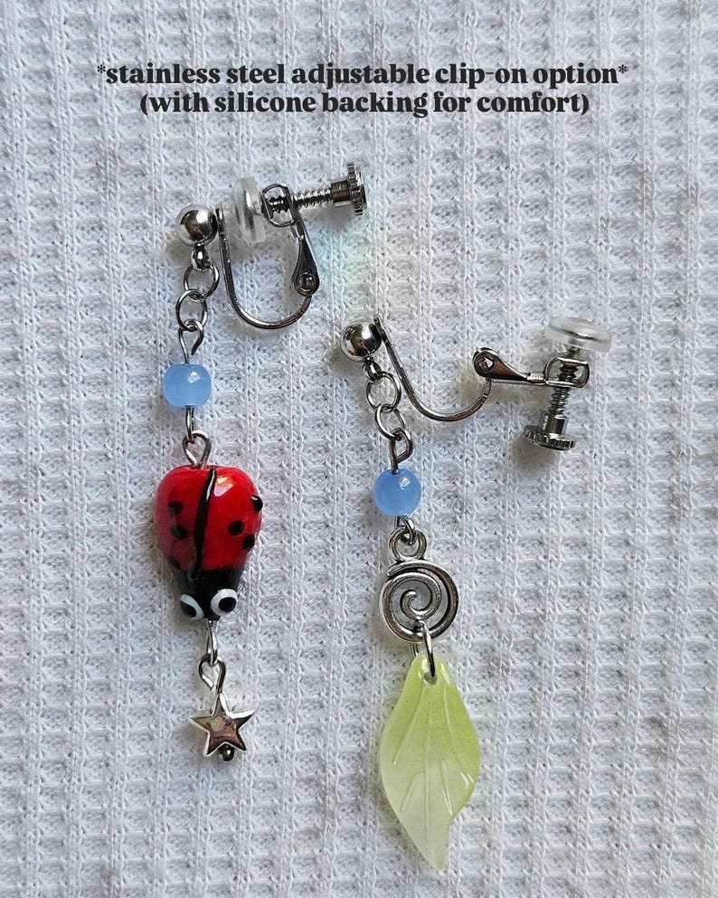 May include: Clip-on earrings with stainless steel adjustable clips. One earring features a red ladybug charm with black spots and a star pendant. The other earring has a green leaf charm and a spiral design. Both have blue beads.