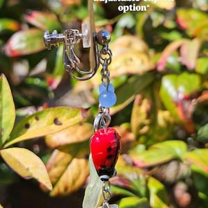May include: A stainless steel clip-on earring with a red ladybug charm, blue beads, and a silver star. The earring has a clear, leaf-shaped accent. Text on the image reads: *stainless steel adjustable clip-on option*.