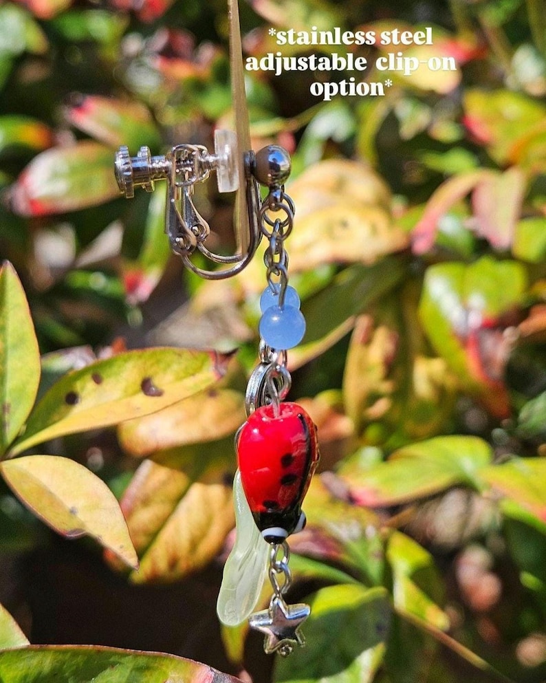May include: A stainless steel clip-on earring with a red ladybug charm, blue beads, and a silver star. The earring has a clear, leaf-shaped accent. Text on the image reads: *stainless steel adjustable clip-on option*.