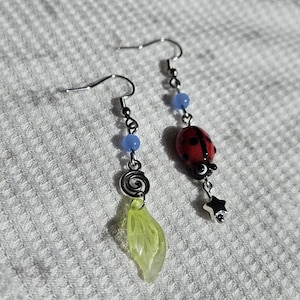 May include: A pair of dangle earrings. One earring features a red ladybug charm with black spots and a silver star. The other earring has a green leaf charm and a silver spiral. Both earrings have blue beads.