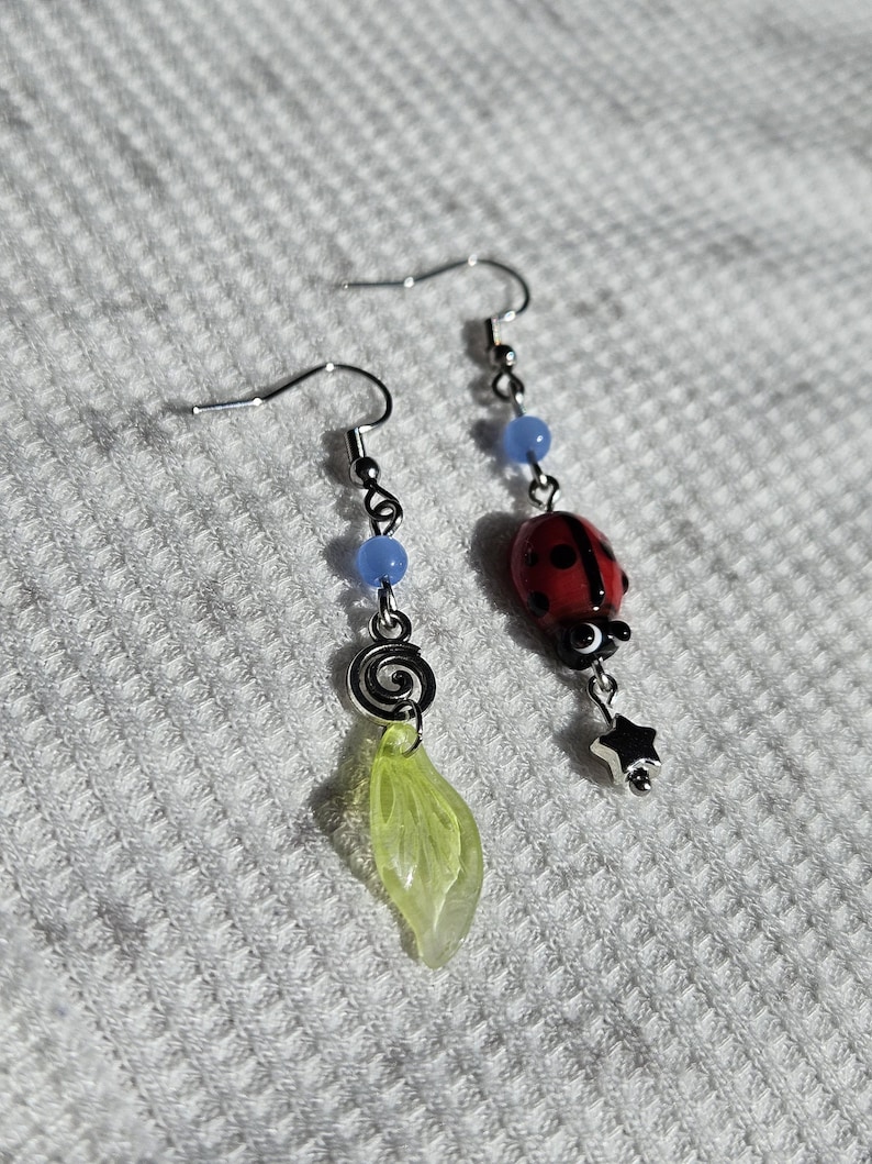 May include: A pair of dangle earrings. One earring features a red ladybug charm with black spots and a silver star. The other earring has a green leaf charm and a silver spiral. Both earrings have blue beads.