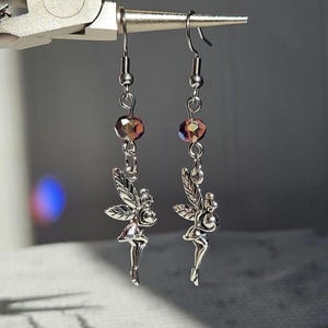 May include: A pair of silver-toned fairy earrings. Each earring features a small fairy charm with detailed wings and a dangling faceted bead in shades of purple and gold. The earrings are suspended from a simple hook.