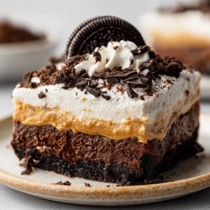 Puede incluir: Una porción de postre en capas sobre un plato beige. El postre tiene una base de chocolate negro, una capa de mantequilla de maní y una cobertura de crema batida. Está adornado con virutas de chocolate y una galleta Oreo.