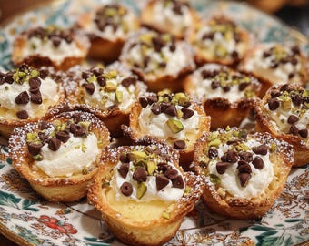 May include: A plate of mini pastries with a golden-brown, flaky crust. Each pastry is filled with a creamy white filling, topped with chocolate chips, chopped pistachios, and a dusting of powdered sugar. The plate has a floral design.