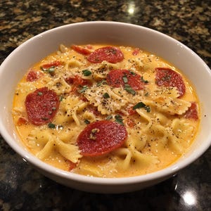May include: A white bowl filled with creamy, orange-colored soup. The soup contains farfalle pasta, red pepperoni slices, and chopped tomatoes. Garnished with herbs and black pepper, this dish is a hearty meal.