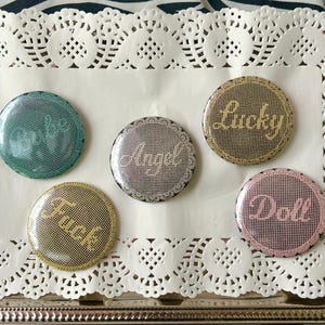 May include: A collection of five round, glittery buttons with various words in cursive script: "Babe", "Angel", "Lucky", "Fuck", and "Doll". The buttons are arranged on a white doily inside a silver tray, set against a zebra-striped background.