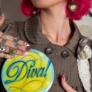 Puede incluir: Una blazer marrón con botones grandes y un lazo decorativo. Se sostiene un letrero redondo amarillo y azul con la palabra «Diva!» frente a la blazer. La persona que sostiene el letrero tiene el pelo rosa, varios anillos y uñas largas decoradas.