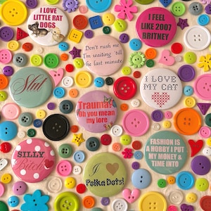 Puede incluir: Una colección de botones y botones de solapa coloridos con varios diseños y texto. Los botones presentan frases como "I love little rat dogs", "I feel like 2007 Britney" y "Fashion is a hobby I put my money & time into."