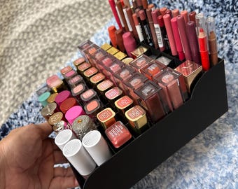 May include: A black organizer filled with various lipsticks, lip glosses, and lip liners in shades of pink, red, and nude. The lipsticks have gold and colored caps. The organizer is held by a hand.