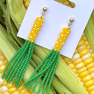May include: A pair of dangle earrings featuring a cylindrical top section made of yellow and white seed beads, attached to a long green seed bead fringe. The earrings are mounted on a white display card with a gold-toned metal post backing.