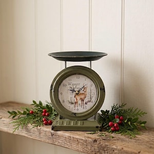May include: An olive green metal scale clock featuring a round face with a deer illustration, the text Winter Wishes, and black hands. The clock has a black circular tray on top and a rectangular base, with a distressed finish throughout.
