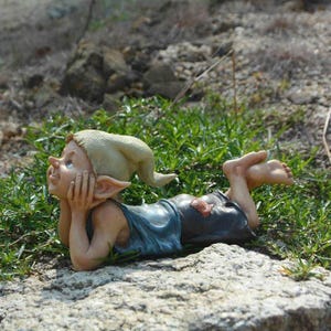 May include: A whimsical garden gnome figurine, lying on a rock and gazing upwards. The gnome wears a blue top, dark shorts, and a pointed hat. The figurine is surrounded by green grass and small rocks, creating a natural outdoor scene.