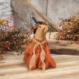 May include: A stuffed fabric mouse figurine wearing a black top hat and a burnt orange dress with a small white dot pattern. The mouse has a tan body, black stitched eyes, and long black thread whiskers, accented by a frayed burlap neck tie.