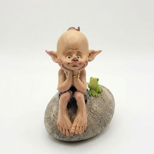 May include: A whimsical figurine of a bald, wide-eyed creature with large ears, seated on a stone. The figure rests its chin on its hands, wearing blue shorts. A small green frog sits nearby. The figurine is a decorative item.