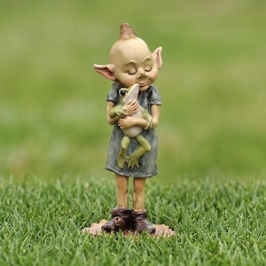 May include: A small figurine of a pointed-eared creature with a bald head, wearing a grey dress and brown boots, holding a light green frog against its chest with eyes closed. The figure stands on a flat, leaf-shaped base with a smooth finish.