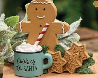 May include: A gingerbread man figurine holding a red and white striped straw, standing next to a blue mug with white marshmallows and the text "Cookies for Santa". Three star-shaped cookies are in the foreground.