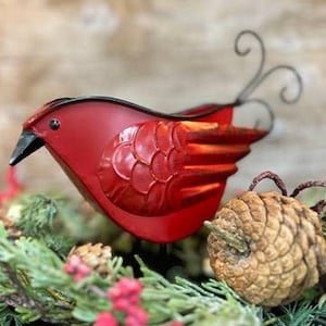 May include: A bright red metal bird figurine with a black beak and a long, curled wire tail. The bird features embossed wing details and is positioned among green pine needles and a brown pinecone, creating a festive, seasonal appearance.