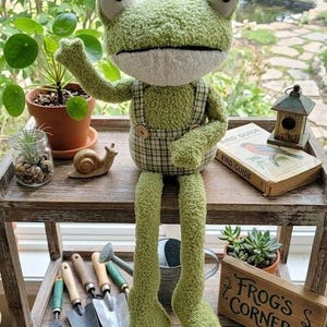 May include: A plush green frog doll with large white eyes and a plaid overall. The frog is sitting on a wooden shelf with gardening tools, potted plants, and a sign that reads "Frog's Corner".