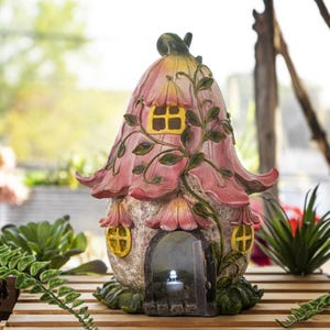 May include: A decorative garden ornament shaped like a house with a pink flower-petal roof, yellow windows, and a textured stone-like base. A small arched doorway features a single illuminated white LED light inside the structure.