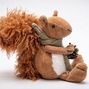 May include: A plush squirrel toy with a brown corduroy body, a white fuzzy belly, and a large, textured, curly brown tail. The squirrel wears a green and white herringbone patterned scarf around its neck and holds a small brown pinecone.