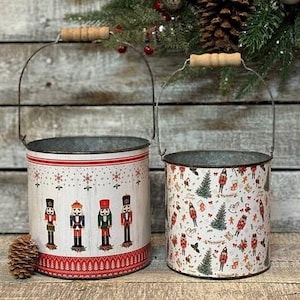 May include: Two cylindrical metal buckets with wire handles and wooden grips. The larger bucket features a row of nutcracker figures and red snowflake patterns. The smaller bucket displays a repeating pattern of nutcrackers, pine trees, and holiday icons.