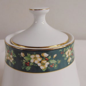 May include: White ceramic sugar bowl with a lid. The bowl features a dark teal band with a floral pattern of white flowers, green leaves, and red berries. Gold trim accents the rim and lid. A classic piece for serving sugar.
