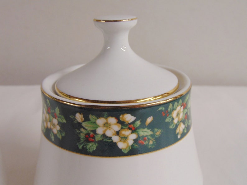 May include: White ceramic sugar bowl with a lid. The bowl features a dark teal band with a floral pattern of white flowers, green leaves, and red berries. Gold trim accents the rim and lid. A classic piece for serving sugar.