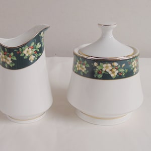 May include: White ceramic creamer and covered sugar bowl set. The set features a dark green band with a floral pattern of white flowers and red berries. Gold trim accents the edges of the pieces.