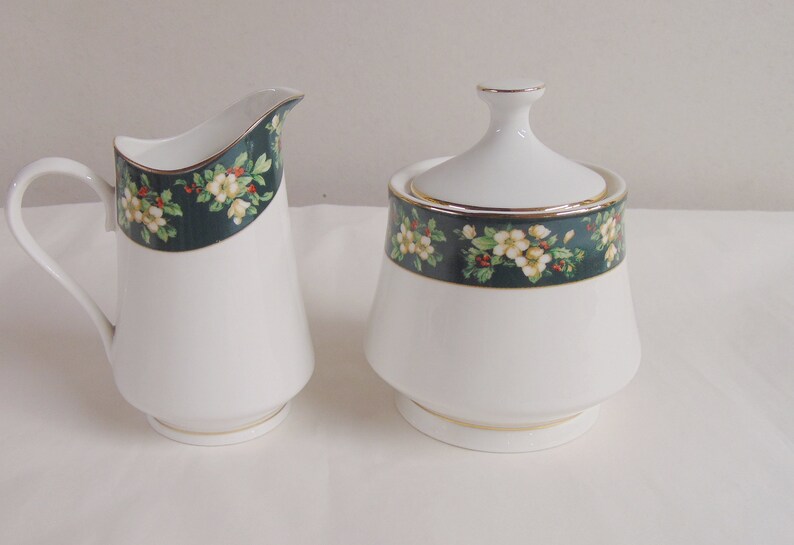 May include: White ceramic creamer and covered sugar bowl set. The set features a dark green band with a floral pattern of white flowers and red berries. Gold trim accents the edges of the pieces.