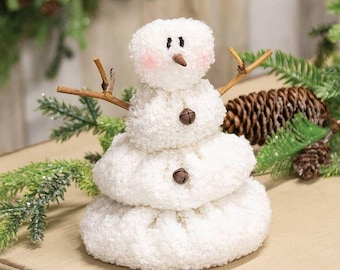 May include: A white, textured, three-tiered snowman figurine made of soft, fuzzy material. It features a small brown carrot-shaped nose, black stitched eyes, pink blush on the cheeks, two brown metal jingle bells as buttons, and twig arms.