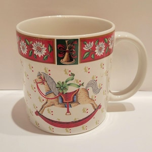 May include: A white ceramic mug with a Christmas theme. The mug features a rocking horse design, red and green accents, and a decorative border with flowers and a bell. The handle is white.
