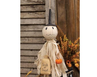 May include: A hanging fabric ghost decoration featuring a beige cloth body with frayed edges, a black top hat, and a small beige sack printed with the text Trick or Treat. A miniature orange pumpkin is attached to the side of the ghost figure.