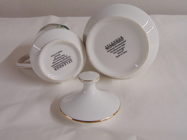 May include: White porcelain creamer and sugar bowl set with gold trim. The creamer has a handle and a floral design. The sugar bowl has a lid with a gold trim. The set is labeled "SPLENDOR" and "Sakura".