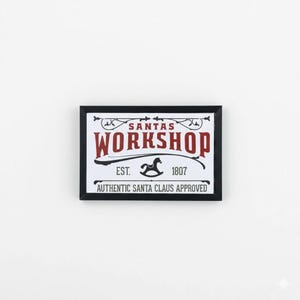 May include: A rectangular sign with a black frame features the text "SANTA'S WORKSHOP" in burgundy, with a rocking horse illustration and the text "EST. 1807." Below, it reads "AUTHENTIC SANTA CLAUS APPROVED."