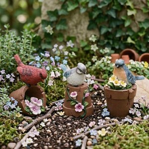 May include: Three miniature bird figurines in a garden setting. A red bird sits atop a broken terracotta pot with pink flowers. A blue bird rests on a decorative pot with floral accents. Another blue bird sits in a pot with yellow flowers.