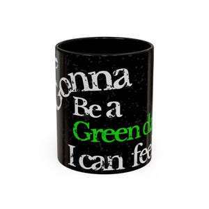 May include: Black ceramic mug with white and green text. The text reads "Gonna Be a Green Day I can feel". The mug has a speckled texture and a glossy finish. The mug is cylindrical and has a handle.