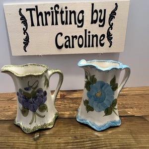 May include: Two ceramic pitchers with floral designs. One pitcher features purple flowers and green leaves, while the other has blue flowers and green leaves. The pitchers have a white base with a scalloped edge. A sign above reads "Thrifting by Caroline."