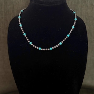 May include: A beaded necklace with alternating silver-colored and turquoise beads. The necklace is displayed on a black mannequin. The beads are small and evenly spaced, creating a delicate and elegant look. The necklace is a jewelry item.