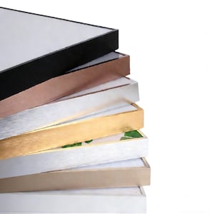 May include: A stack of picture frames with various metallic finishes, including black, copper, silver, gold, and beige. The frames have a clean, modern design with a white interior, showcasing a variety of color options for home decor.