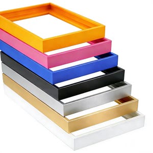 Op de afbeelding: Een stapel rechthoekige fotolijsten in verschillende kleuren: oranje, roze, blauw, zwart, zilver, goud en wit. De lijsten hebben een diep profiel, geschikt voor canvas kunst of ingelijste prints.