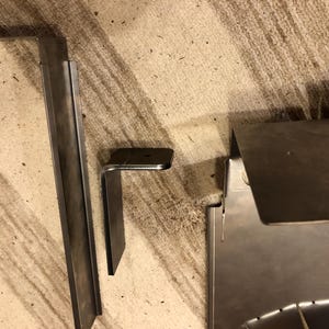 May include: An assortment of unfinished metal components, including a long, rectangular piece, an L-shaped bracket, and a larger, angled sheet. The pieces are likely for construction or assembly purposes, with a raw, industrial aesthetic.