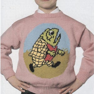 Op de afbeelding: Een lichtroze gebreide trui met een cartoon visontwerp. De vis is geel, groen en rood, tegen een blauwe en bruine achtergrond. De trui heeft een ronde hals en lange mouwen.