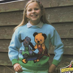 Puede incluir: Una niña lleva un suéter azul claro con un diseño de Sooty y Sweep. El suéter tiene cuello y puños verdes, y la palabra "SOOTY" se muestra en la parte superior. También lleva pantalones azul claro. Colección de personajes.