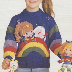 Puede incluir: Un suéter azul tejido con un diseño de arcoíris y personajes de dibujos animados. El suéter tiene cuello alto y mangas a rayas. También se ve una muñeca con pelo amarillo y un peluche a juego.