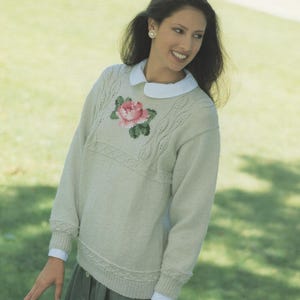 May include: A pale green knitted sweater with a white collar and cuffs. The sweater has a pink and green rose design on the chest. The woman is wearing a skirt and is smiling.