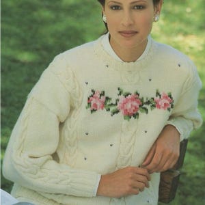 Op de afbeelding: Crèmekleurige kabelgebreide trui met een bloemenontwerp van roze rozen en groene bladeren over de borst. De trui heeft een ronde hals en lange mouwen. Kleine zwarte stippen zijn over de trui verspreid.