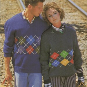 May include: Two people wearing matching argyle sweaters. One sweater is navy blue, the other is dark gray. Both sweaters have a V-neck and feature a colorful argyle pattern across the chest. The person on the left is wearing blue jeans.