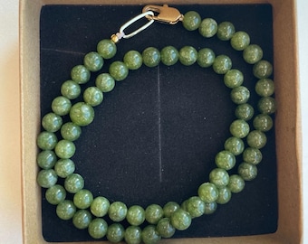 Puede incluir: Un collar de cuentas verdes con un cierre dorado, presentado en una caja marrón con interior negro. El collar está compuesto por cuentas redondas de color verde claro. El cierre es de color dorado.