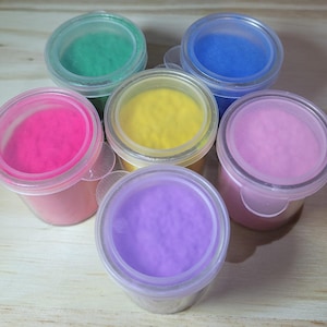 Puede incluir: Seis pequeños recipientes de plástico transparente llenos de polvos de colores. Los colores incluyen verde, azul, rosa, amarillo, morado y rosa claro. Cada recipiente tiene una tapa transparente y está dispuesto en un patrón circular sobre una superficie de madera.