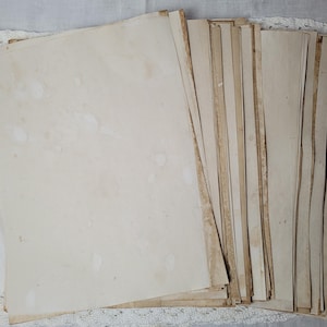 May include: A stack of aged, off-white paper sheets with visible stains and discoloration. The edges of the paper show a darker, aged tone, suggesting a vintage or antique aesthetic. The paper rests on a white lace-trimmed surface.